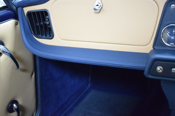 Interior view of a car showing a tan and blue door panel with a vent, glove compartment, and part of a Racetorations Double Stitched Dashboard Top and Crash Pad Cover Set - TR5-6 featuring a clock and button controls.