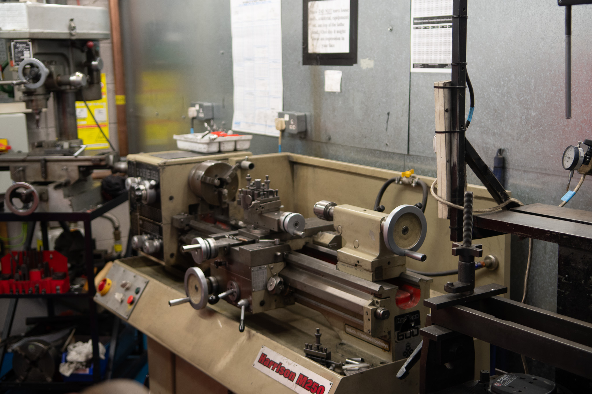 A metal lathe setup in a workshop, surrounded by various tools and equipment. The lathe is equipped with handles, controls, and a chuck holding a cylindrical piece for machining. The workshop walls are covered with metal sheets and some papers and tools hanging.