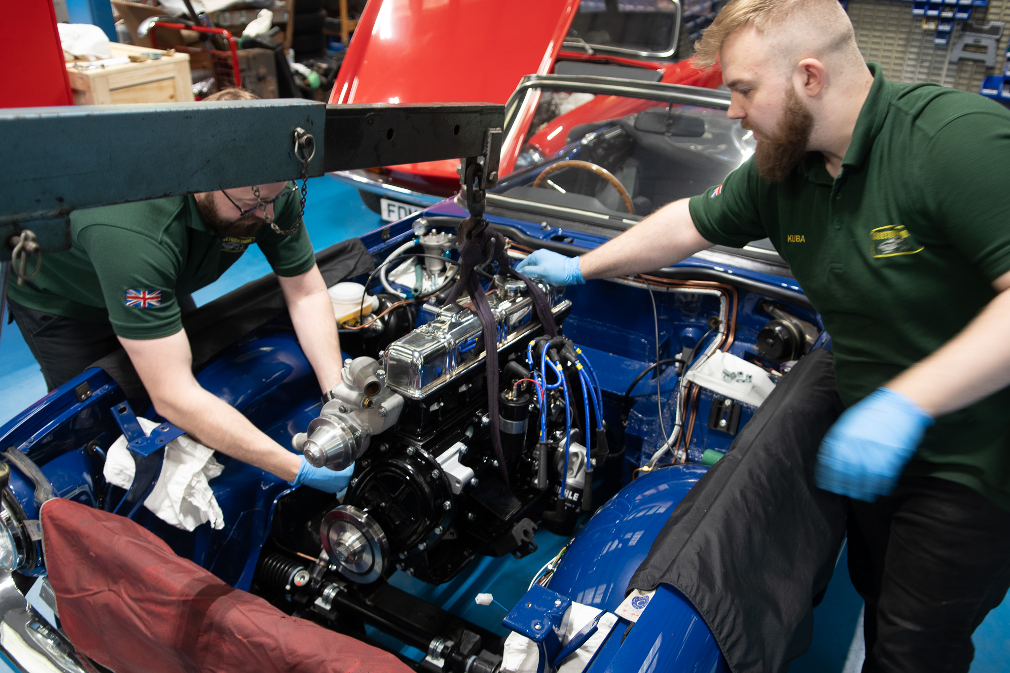 Two mechanics work on installing a car engine. They are in a well-equipped workshop with various tools and parts visible in the background. The car is a blue classic model with the hood open and radiator cover removed. Both mechanics wear uniforms and blue gloves.