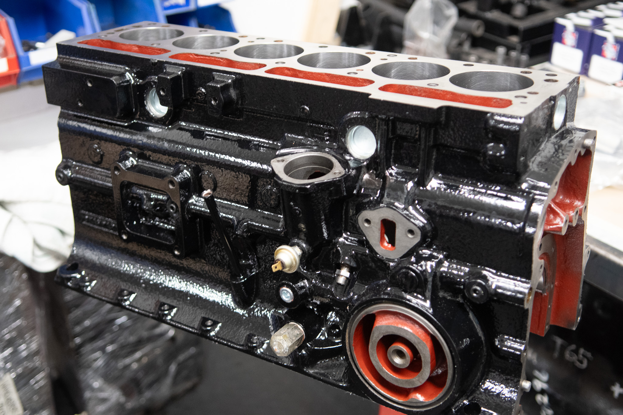 A close-up image of a clean, black and red engine block placed on a workshop bench. The engine block has six cylinders and various components and openings visible. The background shows shelves with tools and mechanical parts.
